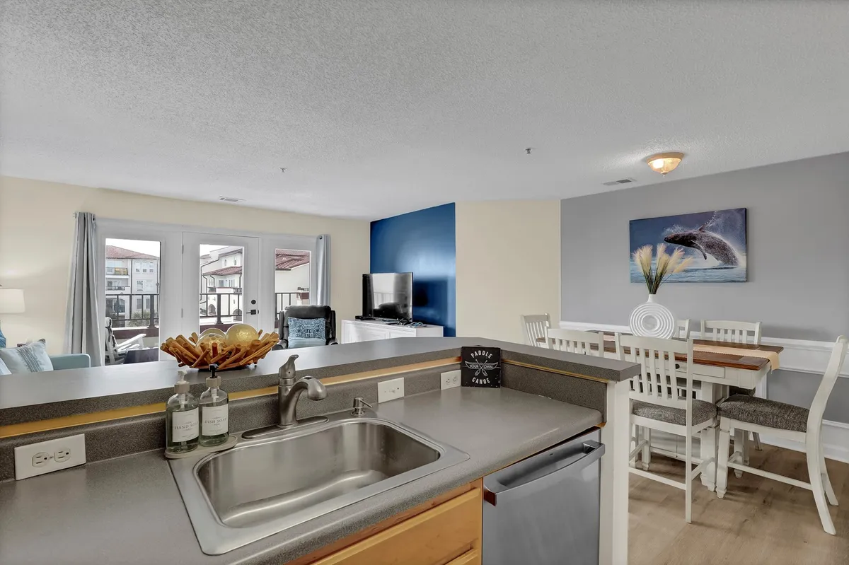 An inviting open-plan living area featuring a kitchen sink, dining table, and a spacious living room. Large windows and a balcony reveal a stunning view, with sunlight streaming in. The decor showcases bold colors and coastal accents, creating a vibrant yet serene atmosphere ideal for relaxation or gatherings.