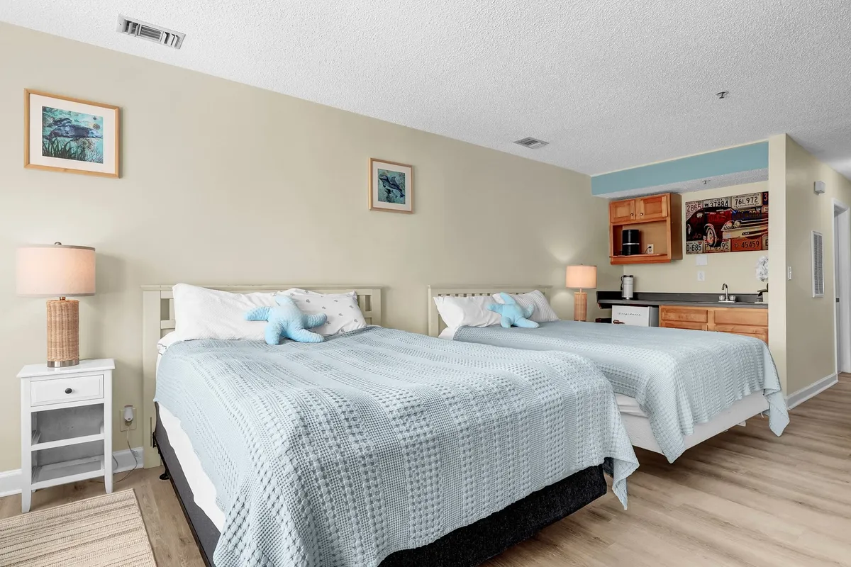A cozy bedroom featuring two beds dressed in light blue blankets, a central nightstand with a softly glowing lamp, and a small kitchenette area. The room exudes comfort and versatility, ideal for friends or families sharing space.