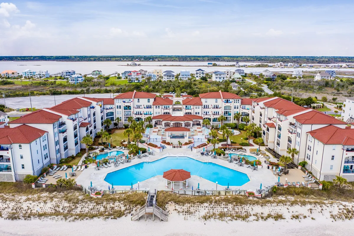 A lively exterior view of a large swimming pool area framed by palm trees, red-roofed resort buildings, and outdoor lounge spaces, with easy access to the beach. Children and families can be imagined playing in the pool, while others relax on lounge chairs under the sun.