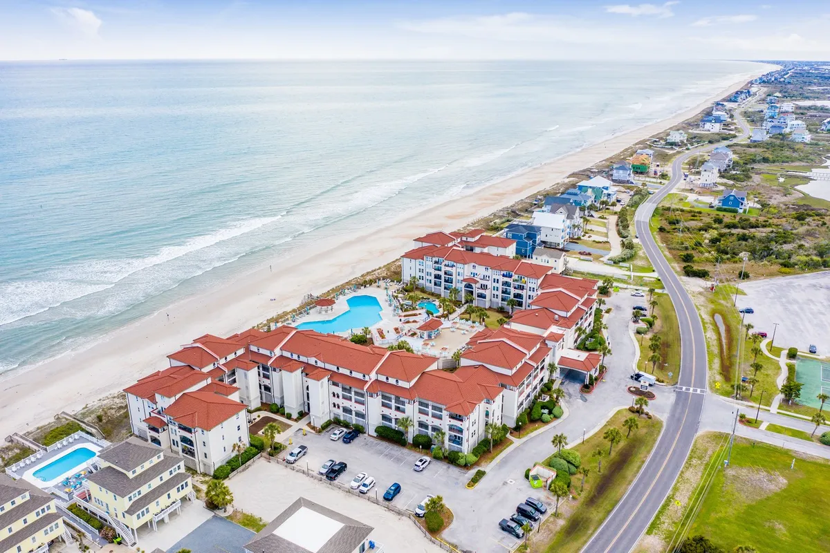 The image captures the inviting pool area of the Villa Capriani complex, set just steps from the soft sands of North Topsail Beach. Sparkling blue water, manicured grounds, and the rolling waves of the Atlantic create a backdrop that invites relaxation and recreation. Guests can envision swimming, sunbathing, and gathering with family or friends, all under the warm North Carolina sun, with ocean views stretching to the horizon.