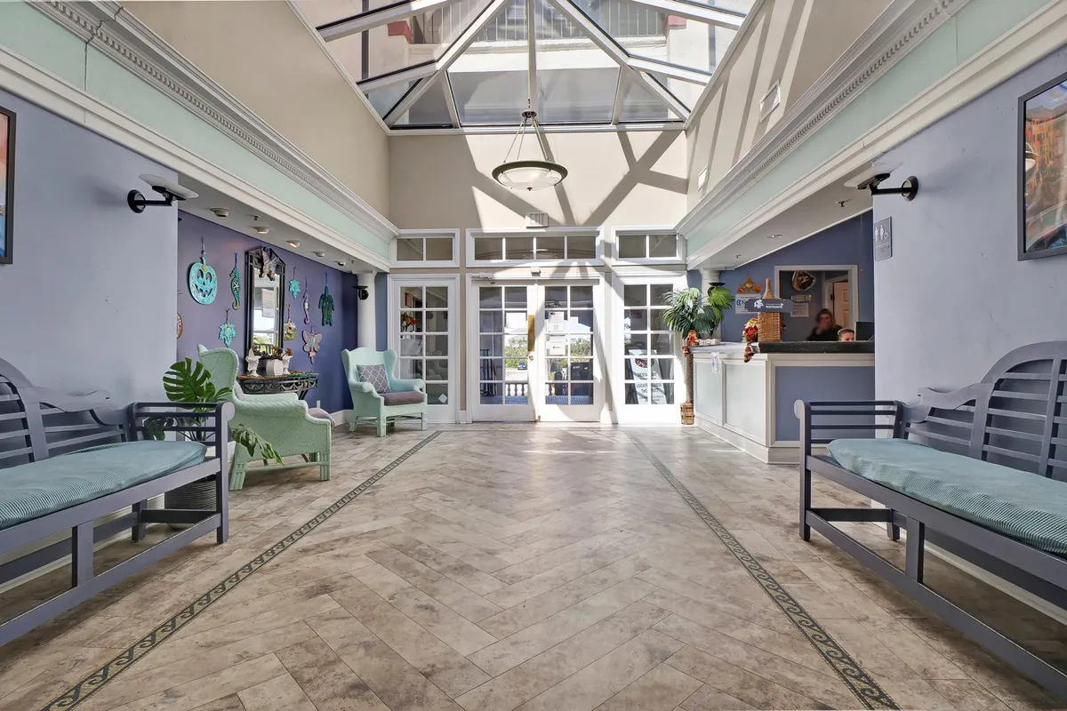 This interior image features a bright and inviting check-in area with a reception desk, green plants, wall art, and a glass ceiling that brings in natural light. Benches offer a welcoming space for guests as they arrive or wait.