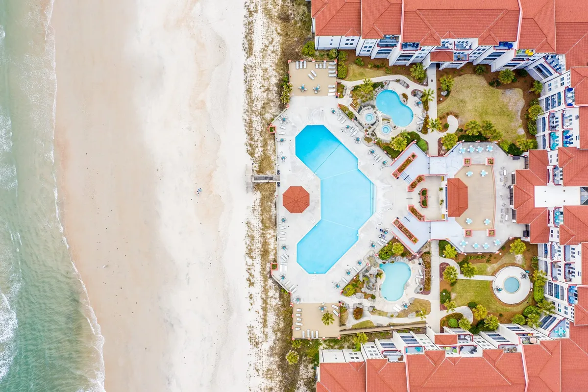 A vibrant scene featuring a large central swimming pool with several smaller pools, all surrounded by red-roofed resort buildings, palm trees, and lounge areas. The area is lively yet relaxed, with direct access to the sandy beach just beyond the pool deck.