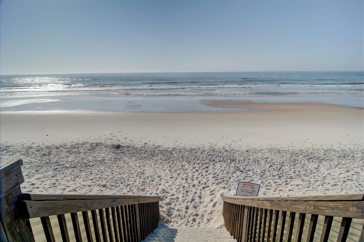 Wide wooden steps descend gracefully to a pristine, quiet shoreline, where gentle ocean waves lap at the sand. The expansive beach invites peaceful strolls and romantic moments at sunrise or sunset.