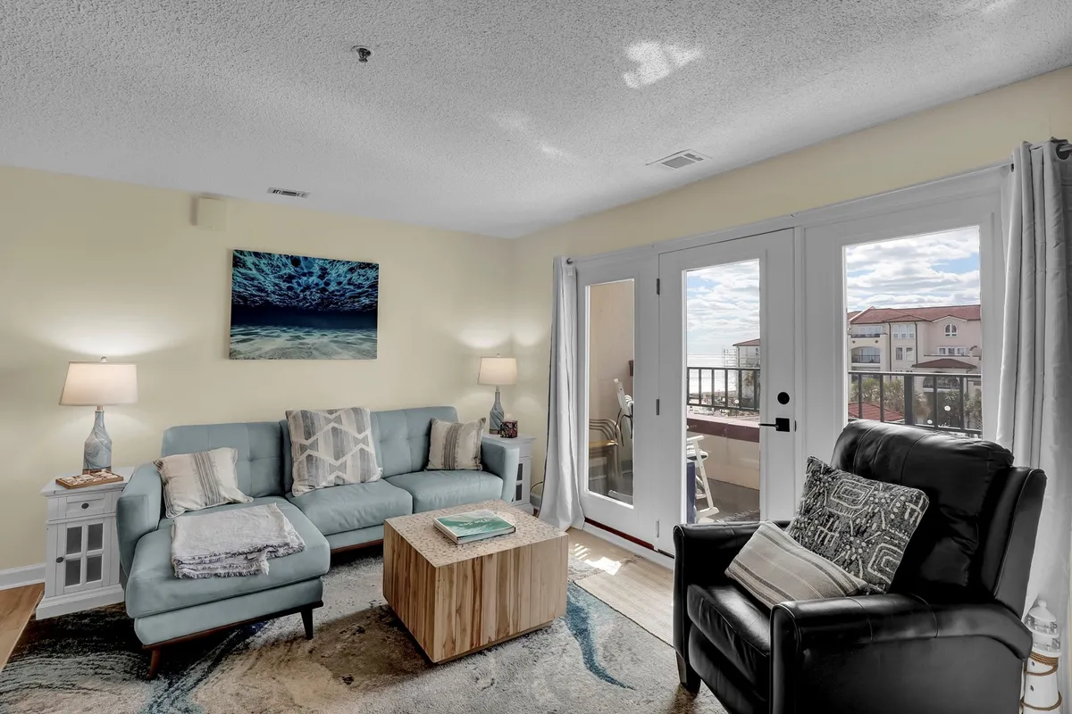 A cozy living space with a light blue sofa, wooden coffee table, large windows letting in natural light, and ocean-themed artwork. The room is styled with soft, calming hues and coastal accents, and the windows provide a glimpse of the terrace or balcony beyond.