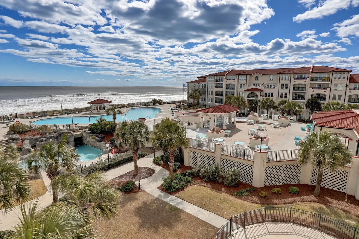 An inviting exterior view showing multiple swimming pools surrounded by landscaped gardens, with balconies overlooking the pool area and the ocean beyond. The tranquil setting is perfect for leisurely swims, sunbathing, or enjoying a sunset from the comfort of your own balcony.