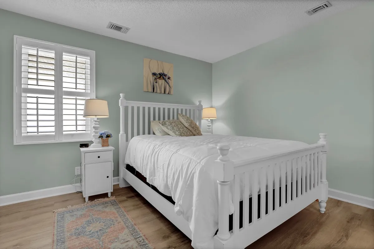 A serene bedroom featuring a crisp white bed with plush decorative pillows and a neatly arranged nightstand with a lamp. Soft artwork hangs above the bed, while natural light pours softly through a nearby window, creating a restful and calming atmosphere.