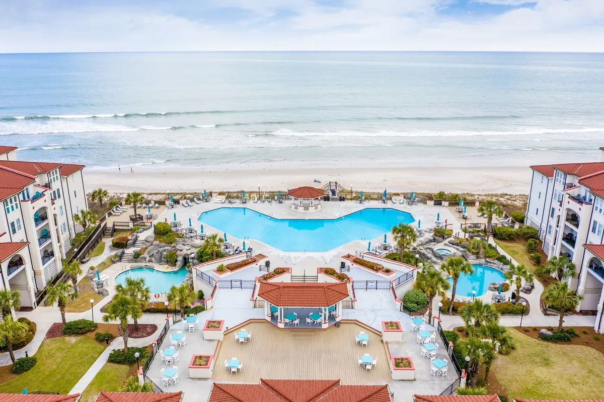 A spacious, resort-style swimming pool surrounded by palm trees, comfortable lounge chairs, and a poolside cabana, all overlooking the ocean and providing direct beach access. The pool deck is bathed in sunlight, offering a tranquil spot for relaxing or social gatherings with views of the shoreline in the distance.