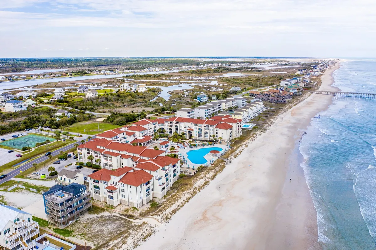 This image offers a stunning exterior view of beachfront properties with a swimming pool, ocean waves rolling in, and a pier reaching into the Atlantic. The scene is bathed in a relaxing light, hinting at sunset and emphasizing the proximity to both water and sand.