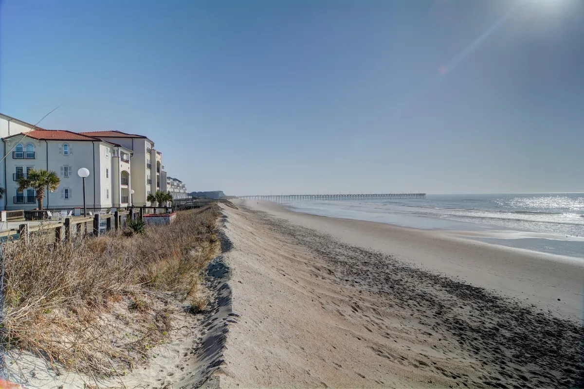 A tranquil beach scene with gentle waves, a long pier stretching into the ocean, and coastal buildings set back from the shoreline. The image captures early morning light and a sweeping stretch of sand ideal for walks or active beach pursuits.