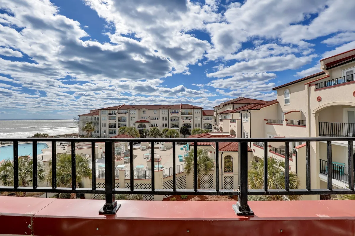 A tranquil balcony scene overlooks a sparkling pool area, with comfortable seating and views of the surrounding buildings and open sky. The setting is designed for relaxation, with space for quiet evenings or social gatherings.
