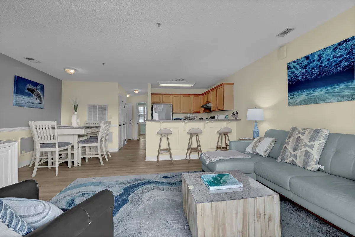 A stylish and inviting living room with a comfortable sofa, dining area, and a kitchen bar with stools in view. Decorative artwork and ample light create a welcoming space for socializing or relaxing, while the sofa hints at the inclusion of a sleeper option for extra guests.