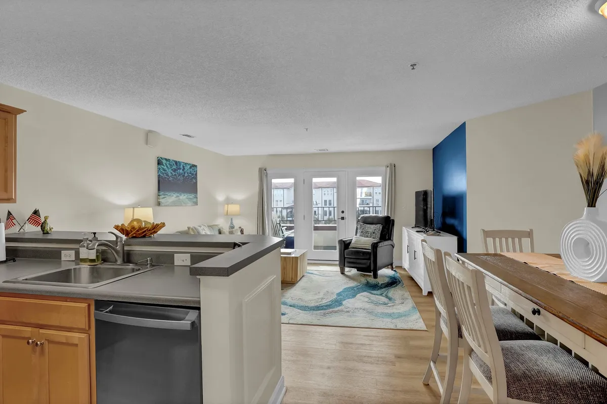 This image shows an open-concept kitchen and dining area with a kitchen counter, adjacent dining table, cozy seating area, and large windows letting in natural light. The scene feels inviting and family-friendly, ideal for shared meals or relaxed gatherings.